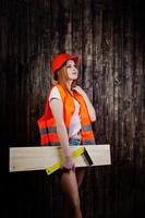 mulher engenheira em laranja protege o capacete e a jaqueta de construção contra o fundo de madeira, segurando a régua e a placa. foto
