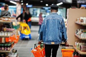 volta do homem afro-americano casual elegante na jaqueta jeans e boina preta segurando duas cestas, andando e fazendo compras no supermercado. foto