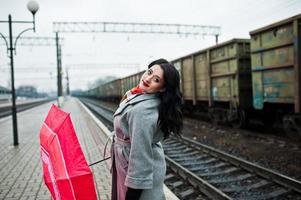 menina morena de casaco cinza com guarda-chuva vermelho na estação ferroviária. foto