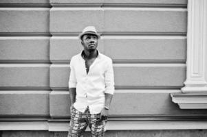homem afro-americano elegante na camisa branca e calça colorida com chapéu posou ao ar livre. menino modelo preto na moda. foto