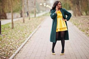 mulher afro-americana elegante no casaco verde e vestido amarelo posou contra o parque outono e falando no telefone. foto