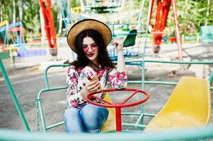 retrato de menina morena de óculos cor de rosa e chapéu com sorvete no parque de diversões. foto