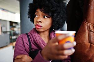 mulher afro-americana de cabelo encaracolado usa suéter posado no café interior com uma xícara de chá ou café. foto