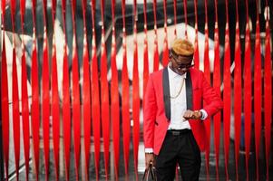 modelo de homem afro-americano de moda no terno vermelho, com destaques de cabelo e bolsa posou na rua e olhando para seus relógios. foto