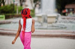 menina afro-americana na moda em calças cor de rosa e dreads vermelhos posou ao ar livre contra fontes. foto