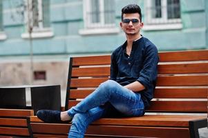 homem elegante modelo indiano em roupas casuais, camisa preta e óculos de sol posou ao ar livre na rua da índia e sentado no banco. foto