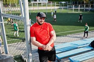 jovem homem musculoso barbudo brutal usar camisa vermelha, shorts e boné no estádio. foto