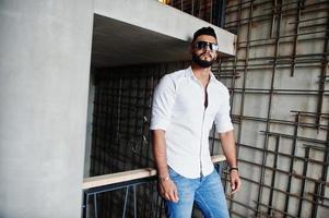 elegante modelo de homem árabe alto na camisa branca, jeans e óculos de sol posou contra a parede de aço interior. barba cara árabe atraente. foto