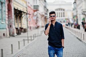 homem elegante modelo indiano em roupas casuais, camisa preta e óculos de sol posou ao ar livre na rua da índia e falando ao telefone. foto