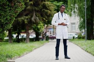 elegante médico americano africano com estetoscópio e jaleco, em óculos posados ao ar livre. foto