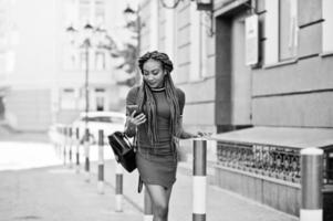 linda e magra garota afro-americana de vestido vermelho com dreadlocks e mochila posou ao ar livre e olhando para o celular na rua. elegante modelo preto. foto