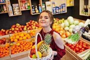 garota de vermelho segurando diferentes frutas e legumes na cesta na loja de frutas. foto