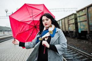 menina morena de casaco cinza com guarda-chuva vermelho na estação ferroviária. foto