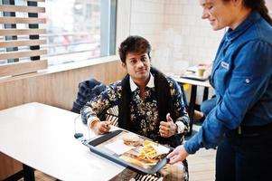 Garçom de garota envia a bandeja com hambúrguer de pedido e batatas fritas para homem indiano elegante atraente e bonito usar camisa no café lounge moderno em seu intervalo de trabalho. foto