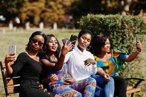 grupo de quatro garotas afro-americanas sentadas no banco ao ar livre com telefones celulares nas mãos e fazendo selfie. foto