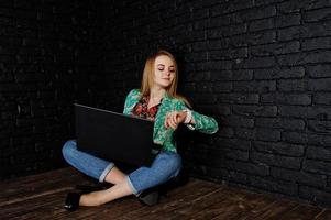 menina loira elegante jaqueta e jeans com laptop olhando relógios contra a parede de tijolo preto no estúdio. foto