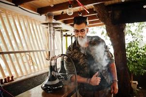 homem árabe de barba elegante de óculos e jaqueta militar fumando cachimbo de água no bar da rua. modelo árabe descansando. foto