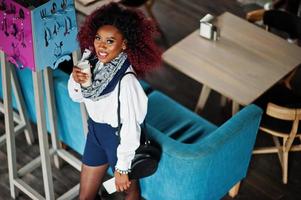 garota encaracolada afro-americana atraente na blusa branca e shorts azuis posou no café com café com leite e celular na mão. vista de cima. foto