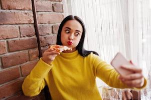 menina morena engraçada de suéter amarelo comendo pizza no restaurante e fazendo selfie. foto