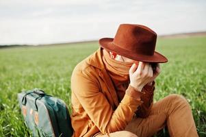 homem elegante de óculos, jaqueta marrom e chapéu com bolsa posou em campo verde. foto