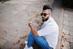 elegante modelo de homem árabe alto na camisa branca, jeans e óculos de sol, sentado na rua da cidade. barba cara árabe atraente no parque. foto