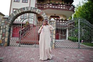rico homem de negócios árabe do Oriente Médio com óculos de sol posou na rua contra a mansão, falando no celular. foto