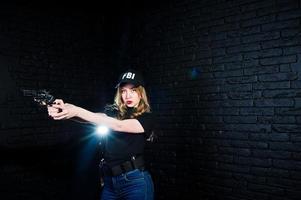 agente feminina do FBI no boné e com arma no estúdio contra a parede de tijolos escuros. foto