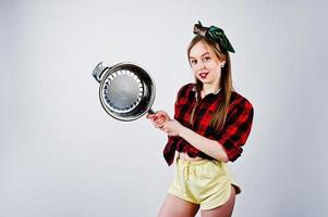 jovem dona de casa engraçada na camisa quadriculada e short amarelo pin up estilo com frigideira isolada no fundo branco. foto