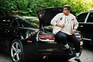 cara de homem árabe elegante hipster posou ao ar livre na rua e sentado no porta-malas seu muscle car preto. homem negro rico. foto