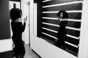 elegante jovem bela mulher afro-americana com penteado afro e hangbag posando desgaste em preto elegante. fazendo foto no espelho por telefone.