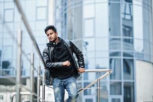 homem asiático elegante e casual na jaqueta de couro preta posou perto da grade contra o edifício moderno. foto