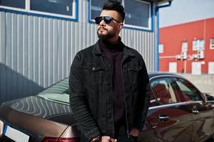 moda homem árabe usar jaqueta jeans preta e óculos escuros posou contra carro moderno de negócios. cara modelo árabe elegante, bem sucedido e elegante. foto