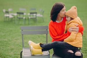 tiro horizontal de mulher carinhosa vestida de suéter de malha vermelha, mantém a filha pequena de joelhos, beija-a na testa, senta-se na cadeira, desfruta de um bom descanso, sente apoio e amor. conceito de família foto