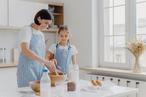 tiro horizontal de mãe e filha carinhosas cozinham juntas, mãe dá aula de culinária para menina, usa farinha, ovos e leite para preparar uma refeição saborosa. tempo para a família, panificação e conceito culinário foto