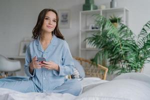 foto interna de mulher jovem e bonita assiste a série favorita com fones de ouvido e telefone inteligente, baixa novas músicas, olha pensativamente de lado, senta-se na cama em um quarto aconchegante. hora de dormir e conceito de lazer