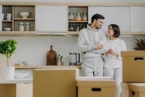 foto interna de um adorável casal recém-casado conversando um com o outro com expressões satisfeitas, bebendo café para viagem em copos de papel, posando na cozinha moderna com móveis novos comprados, mudando de lugar de vida