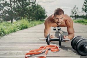 homem bonito motivado forte fica em pose de prancha, faz exercícios abdominais, posa perto de equipamentos esportivos, ouve música em fones de ouvido sem fio, faz exercícios ao ar livre, parece com expressão séria foto