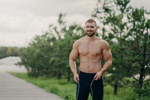 alegre esportista com barba pula com corda, tem corpo musculoso, gosta de treinamento físico, posa ao ar livre. homem forte com torso nu usa equipamentos esportivos para treino, respira ar fresco na natureza foto