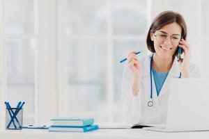 foto de terapeuta ou médico feliz conversa por telefone com o paciente, dá conselhos sobre como curar doenças, usa vestido branco, lê informações no computador portátil, trabalha em clínica privada