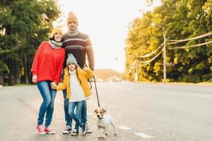 família feliz usa roupas quentes anda com cachorro na estrada, fica perto um do outro como pose na câmera. menina mostra sinal de ok como estar feliz em passar o tempo livre com seus pais e animal de estimação favorito foto