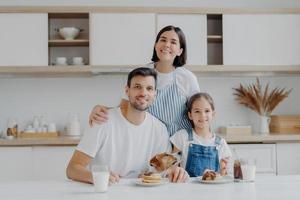 retrato de família de feliz mãe, filha e pai posam na cozinha durante o café da manhã, comem deliciosas panquecas caseiras, seu cachorro posa perto, têm bons relacionamentos amigáveis, se amam foto