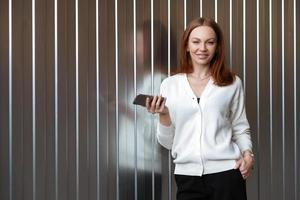 foto de atraente empresária de cabelos castanhos usa roupas formais em preto e branco, usa aplicativo no smartphone, tem maquiagem e manicure, mantém a mão no bolso, fica contra a parede com espaço em branco