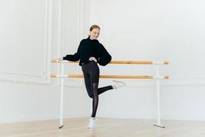 pessoas, dança, coreografia, conceito de flexibilidade. mulher ruiva magra e encantada em traje preto, levanta uma perna, fica perto da barra de balé no estúdio de dança vazio, pratica antes da apresentação. foto