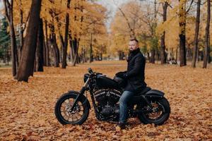 tiro ao ar livre de motociclista masculino com barba grossa, usa luvas de proteção, casaco preto e jeans, posa na moto no belo parque com folhas de laranja, segura o capacete, pára para descansar. estilo de vida urbano foto