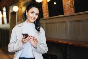 mulher morena bonita com sobrancelhas finas e bem formadas e olhos quentes, vestindo roupas elegantes, sentado no café esperando seu pedido, ouvindo livro de áudio on-line usando o celular tendo sonhos foto