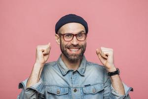 homem barbudo feliz com olhos azuis e expressão alegre cerra os punhos, sente-se vencedor, demonstra positividade, isolada sobre fundo rosa. jovem emocional animado posa no interior foto