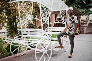 homem afro-americano bonito vestindo roupas tradicionais, boné e óculos perto de carruagem branca. foto