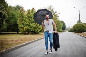 homem de barba árabe alto na moda usar camisa, jeans e óculos de sol andando no parque com guarda-chuva e casaco na mão. foto