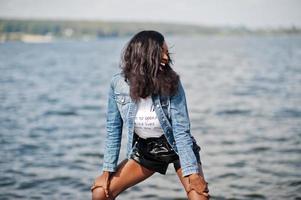 modelo magro de pele escura afro-americana posou em um short preto e jaqueta jeans contra o lado do mar. foto