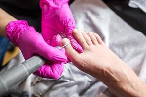 mestre pedicure em luvas cor de rosa corta as unhas da cutícula e goma-laca no salão de pedicure usando broca. pedicure profissional na clínica de cosmetologia. higiene para os pés no salão de beleza. foto